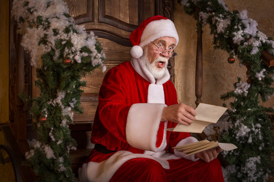 Santa Claus is reading the letters on the porch of decorated house.