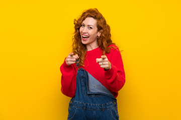 Redhead woman with overalls over isolated yellow wall points finger at you