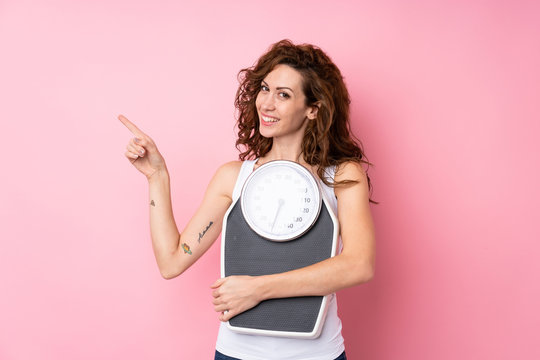 Young woman with curly hair holding a weighing machine over isolated pink background - a woman holdi