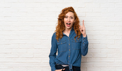 Redhead woman over white brick wall thinking an idea pointing the finger up