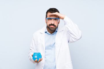 Young scientific holding laboratory flask over isolated background looking far away with hand to...