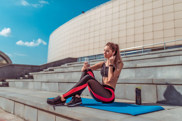 Girl athlete sits mat yoga gymnastics, phone listens music headphones, rest workout, summer day city. Enjoys listens podcast lecture on Internet. Active lifestyle fitness, fashionable stylish woman.