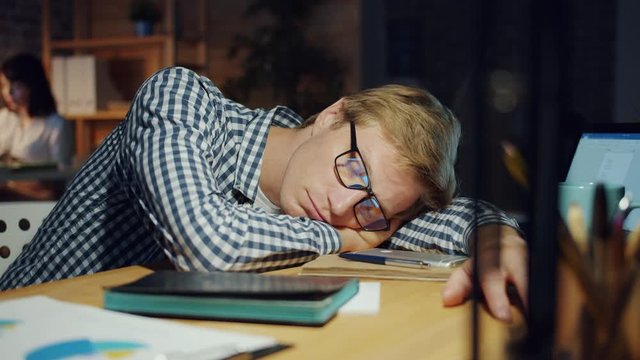 Slow motion of handsome guy in glasses sleeping on table in dark office relaxing at work feeling tired exhausted. People, lifestyle and workplace concept.