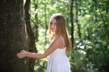 Girl leaning on a tree in a forest in a white dress and looking