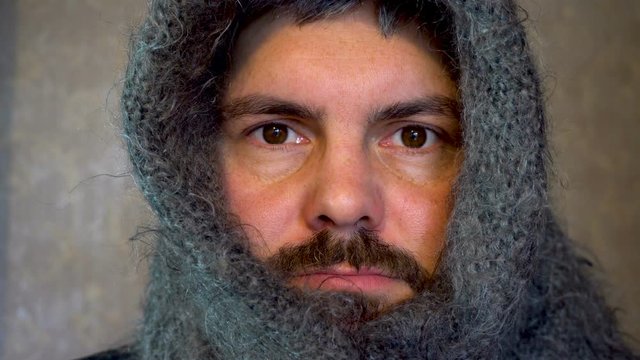 A Man In A Warm Shawl On His Head. A Bearded Man Has A Sick Look. Blinks Eyes. Against The Background Of Gray Wallpaper On The Wall.