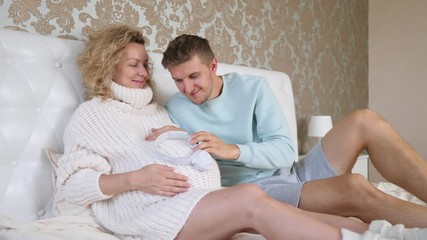 Portrait Of Young Happy Couple At Home. Future Father Putting Headphones On His Pregnant Wife's Belly. - Powered by Adobe