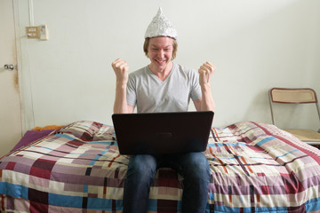 Happy young man with tin foil hat using laptop and getting good news in the bedroom