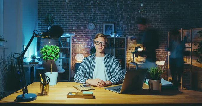 Time lapse of blond guy businessman sitting at desk in office late at night looking at camera, laptop computer is visible on table. Colleageus are rushing in background.