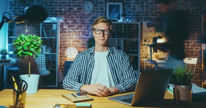 Time lapse of attractive blond guy in glasses looking at camera in illuminated office late night sitting at desk while coworkers are rushing in background.