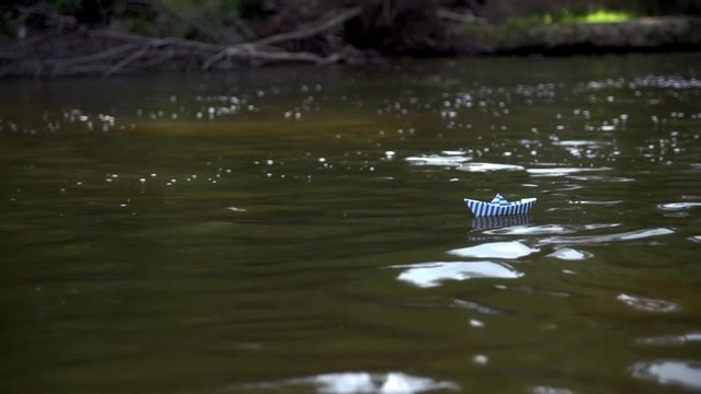 Striped in blue and white stripes paper boat floats in dirty water of river evoking feelings and emotions of powerlessness and defencelessness.