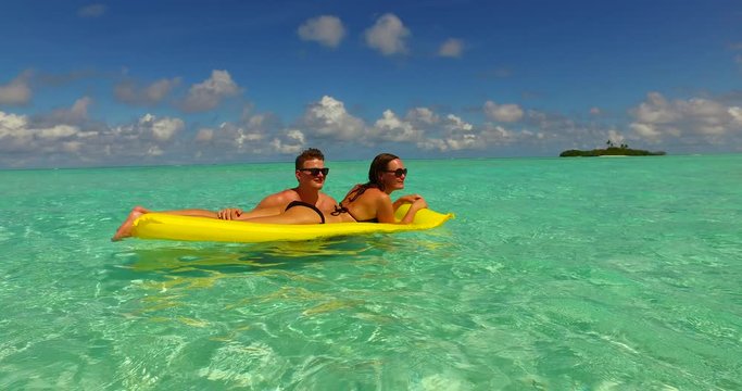 Couple relaxing off the shore of tiny tropical island on yellow inflatable, with girl wearing sunglasses and black bikini