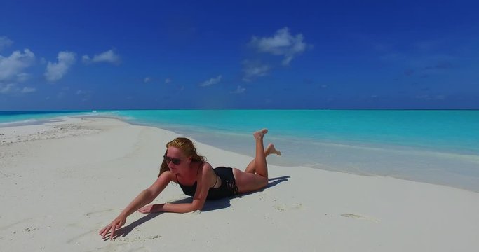 A young woman wearing black sunglasses and a black swim suit relaxes on a tropical shore and draws in the sand in the Maldives