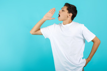 Cheerful young brunet man in white t-shirt is holding hand near her open mouth on blue background