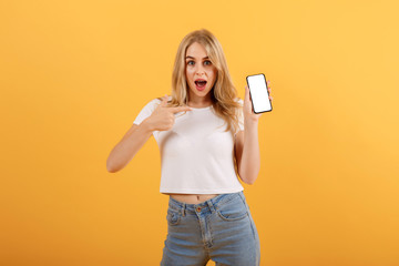 Surprised girl in white t-shirt is on a orange background, pointing her finger at the smartphone in her hand