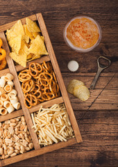 Glass of craft lager beer and opener with box of snacks on wooden background. Pretzel,salty potato sticks, peanuts, onion rings with nachos in vintage box with openers and beer mats. Top view