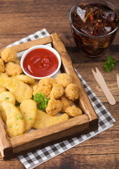 Buttered chicken nuggets and popcorn bites in vintage wooden box with ketchup and glass of cola on wooden background.