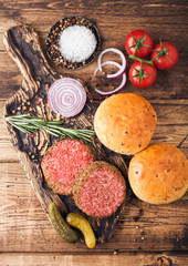 Fresh raw minced pepper beef burgers on vintage chopping board with buns onion and tomatoes on wooden background. Top view