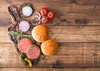 Fresh raw minced pepper beef burgers on vintage chopping board with buns onion and tomatoes on wooden background. Space for text
