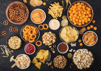 All classic potato snacks with peanuts, popcorn and onion rings and salted pretzels in bowl plates on black background. Twirls with sticks and potato chips and crisps with nachos and cheese balls.