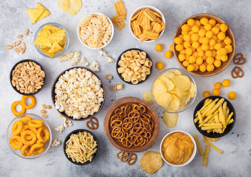 All classic potato snacks with peanuts, popcorn and onion rings and salted pretzels in bowl plates on light background. Twirls with sticks and potato chips and crisps with nachos and cheese balls.