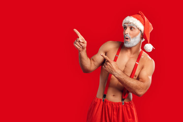 Fototapeta na wymiar Christmas Freestyle. Young bearded Santa Claus bare muscular upper body in hat standing isolated on red pointing aside at copy space excited