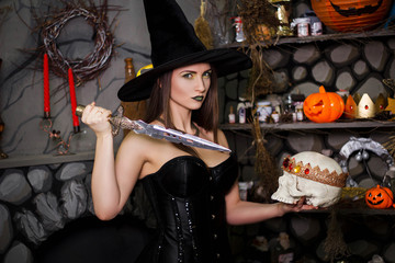 A young, beautiful girl in a green skirt, a black corset and a black hat holds a knife and a skull with a crown in her hands. Witch costume, Halloween, photo studio, model with clean skin.