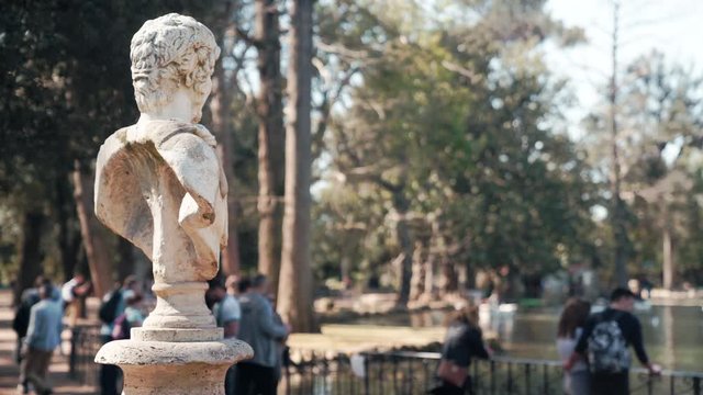 Historic italian marble statue of ancient roman emperor on the backgroundof crowded city park. People walking in the park and taking pictures of ancient lake. Famous roman monument