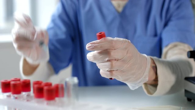 Close up shot of male scientist in sterile gloves drawing blood with syringe from test tube while working at desk in laboratory