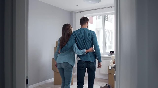Back view of attractive couple in love which embracing and approaching to the big window in their new flat