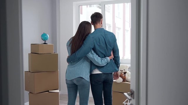 Back view of adorable happy couple which standing embracing in the middle of the empty new flat in front of window