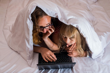 Mom and little daughter lying in bed under a blanket and looking at a laptop.