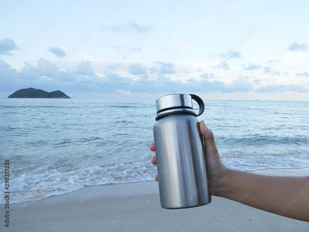 Wall mural close up hand holding reusable water bottle with sea and sand background. save the world and sea lif