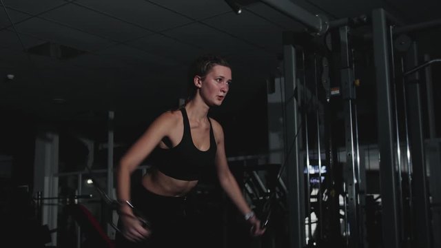 Steadicam shot of a slender pretty Caucasian girl training the muscles of the shoulders and back on a multifunctional simulator in a dark room of the gym. Motivation to sport, healthy lifestyle