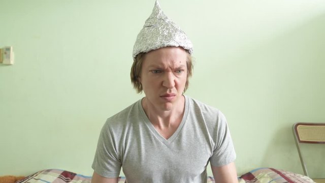Face of stressed young man with tin foil hat having headache in the bedroom