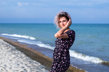 Fototapeta na wymiar beautiful female with curly hairs in black dress in flowers is standing near water on the beach. Orange sunset light on the face