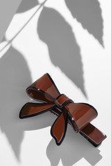 Subject shot of a cambered hair clip made of burgundy glazy material in the form of a bow-knot with a black stripe. The image is made in the studio on the white surface with shadows of the leaves. © RedUmbrella&Donkey