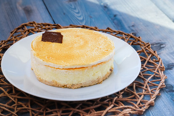 On a dark wooden tabletop on chocolate-colored round wicker straw napkin on a white dish, a round cheesecake with chocolate on the cake.
