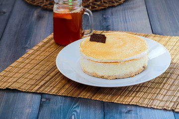 On a dark wooden tabletop on a straw napkin on a round white dish, cake cheesecake and tea with lemon in a glass mug with a green straw.
