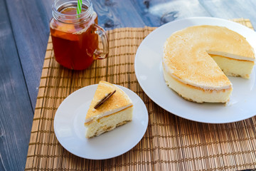 Classic cheesecake cake on a dish, a piece of cake on a saucer, a spoon, tea in a mug, everything on a straw napkin on a dark wood tabletop