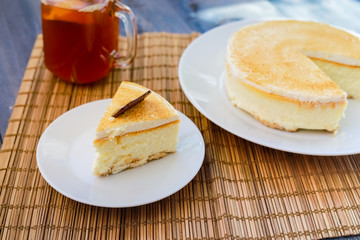 Classic cheesecake cake on a dish, a piece of cake on a saucer, a spoon, tea in a mug, everything on a straw napkin on a dark wood tabletop