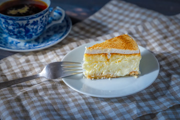 On a dark countertop, on a light checkered linen napkin, a white and blue patterned tea pair with tea and saucer with a piece of classic cheesecake and a fork.