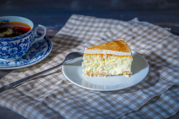 On a dark countertop, on a light checkered linen napkin, a white and blue patterned tea pair with tea and saucer with a piece of classic cheesecake and a fork.