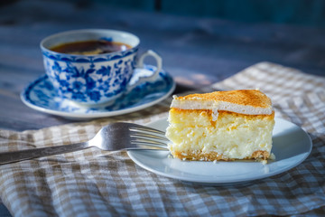 On a dark countertop, on a light checkered linen napkin, a white and blue patterned tea pair with tea and saucer with a piece of classic cheesecake and a fork.