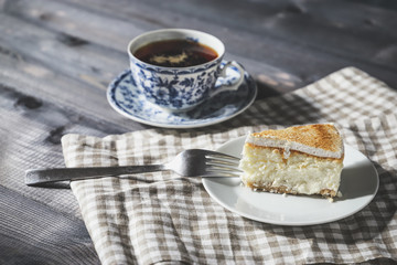 On a dark countertop, on a light checkered linen napkin, a white and blue patterned tea pair with tea and saucer with a piece of classic cheesecake and a fork.
