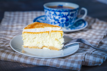 On a dark countertop, on a light checkered linen napkin, a white and blue patterned tea pair with tea and saucer with a piece of classic cheesecake and a fork.