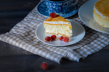 On a dark tabletop, on a light checkered linen napkin on a white saucer, a piece of cheesecake cake and a tea pair with tea