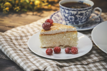 On the saucer is a piece of cheesecake cake and red berries, the whole cake on a white plate, next to it is a white tea pair with a blue pattern, everything is on a linen napkin.