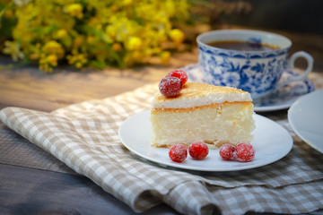 On the saucer is a piece of cheesecake cake and red berries, the whole cake on a white plate, next to it is a white tea pair with a blue pattern, everything is on a linen napkin.