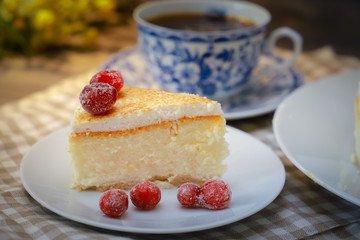 On the saucer is a piece of cheesecake cake and red berries, the whole cake on a white plate, next to it is a white tea pair with a blue pattern, everything is on a linen napkin.