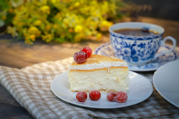 On the saucer is a piece of cheesecake cake and red berries, the whole cake on a white plate, next to it is a white tea pair with a blue pattern, everything is on a linen napkin.